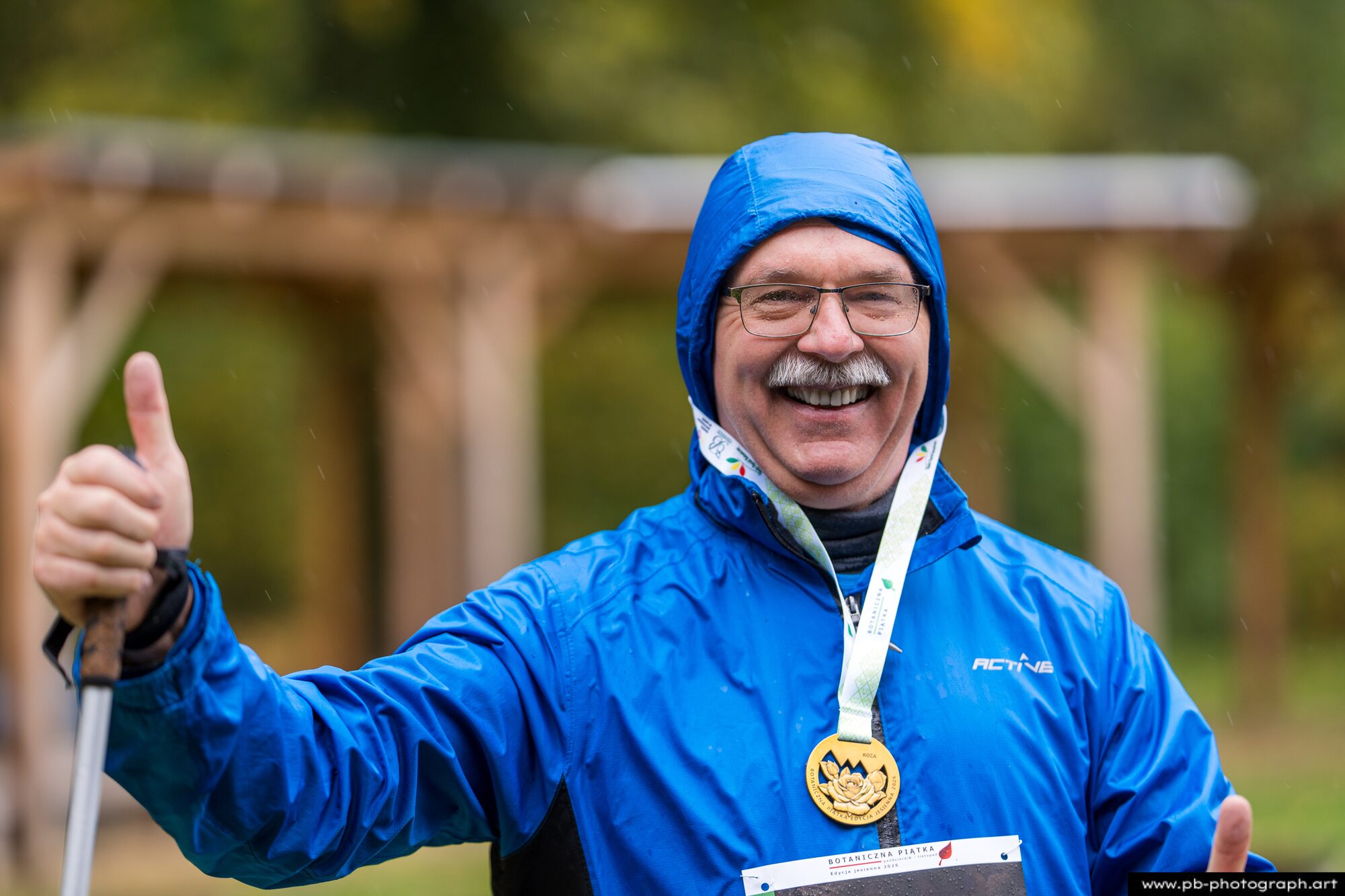 Uśmiechnięty uczestnik w niebieskiej kurtce z medalem pokazuje kciuk po ukończeniu biegu